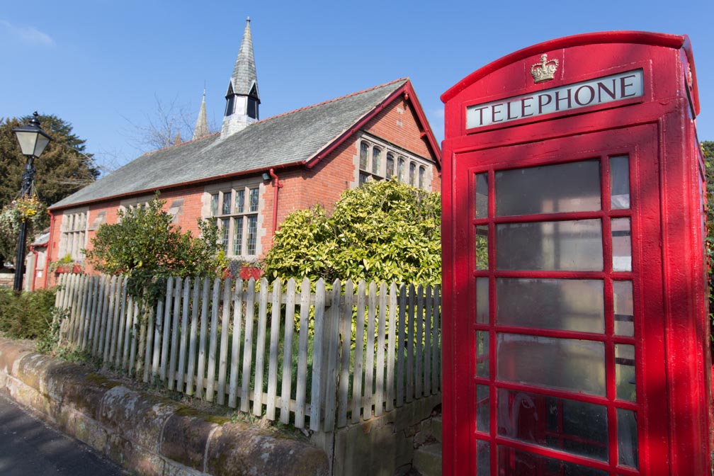 Village of Aldford, England. Picturesque view of Grade II listed K5 red telephone box with Aldford Village Hall in the background.